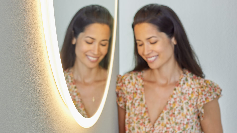 Woman smiling in front of a Geberit Option round mirror with floating design, available in four sizes