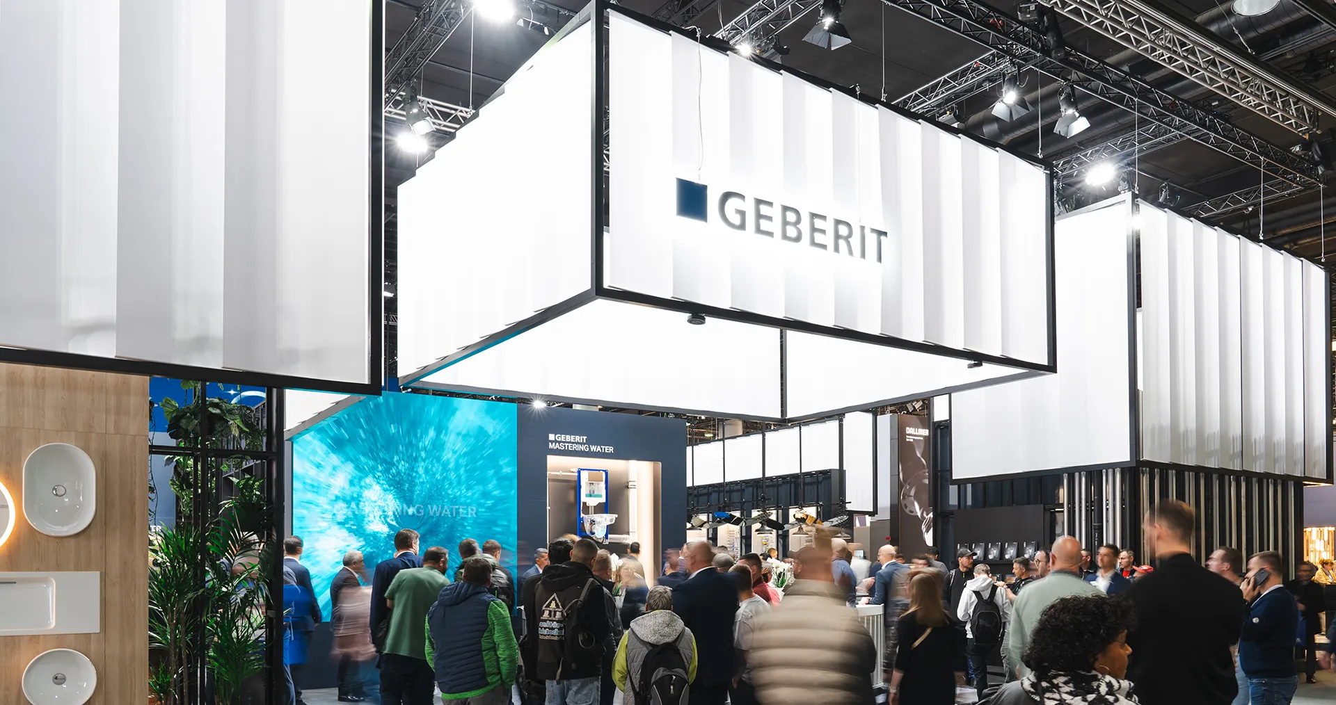 The image shows an exhibition with a modern and geometric architectural design. There are people gathered around booths and information stands. There's a clear sign that says "Geberit", the company being represented at one of the booths. (Photo)