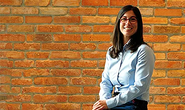 Ágnes Farkas is wearing glasses and a blue blouse and is smiling at the camera in front of a wall. (Photo)