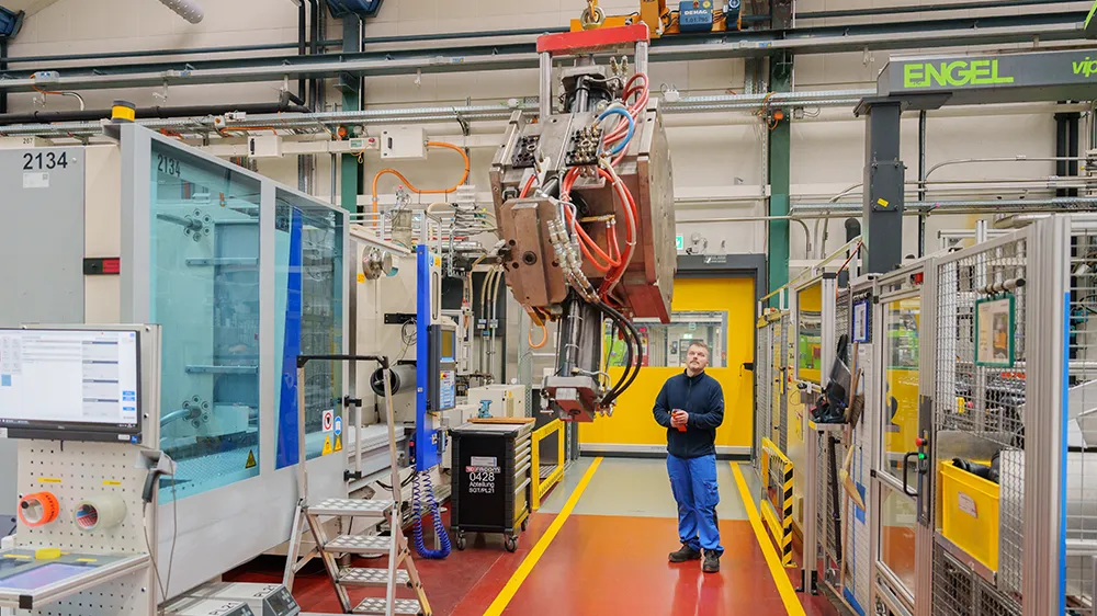 An injection moulding machine must be retooled according to plan and equipped with a new injection mould. Using the indoor crane, Tobias Höhn carefully lifts the mould, which weighs several tonnes, into the air. (Photo)