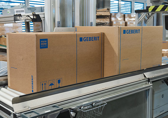 Cardboard boxes with the Geberit logo on a conveyor belt, indicating packaged products ready for distribution