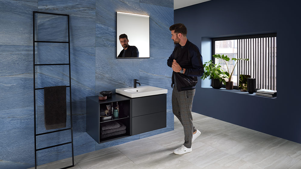 Mann reflektiert auf Geberit ONE Waschtischmöbel und Geberit Option Square Spiegel mit mattschwarzem Rahmen in einem stilvollen Badezimmer mit blauen Steinfliesen