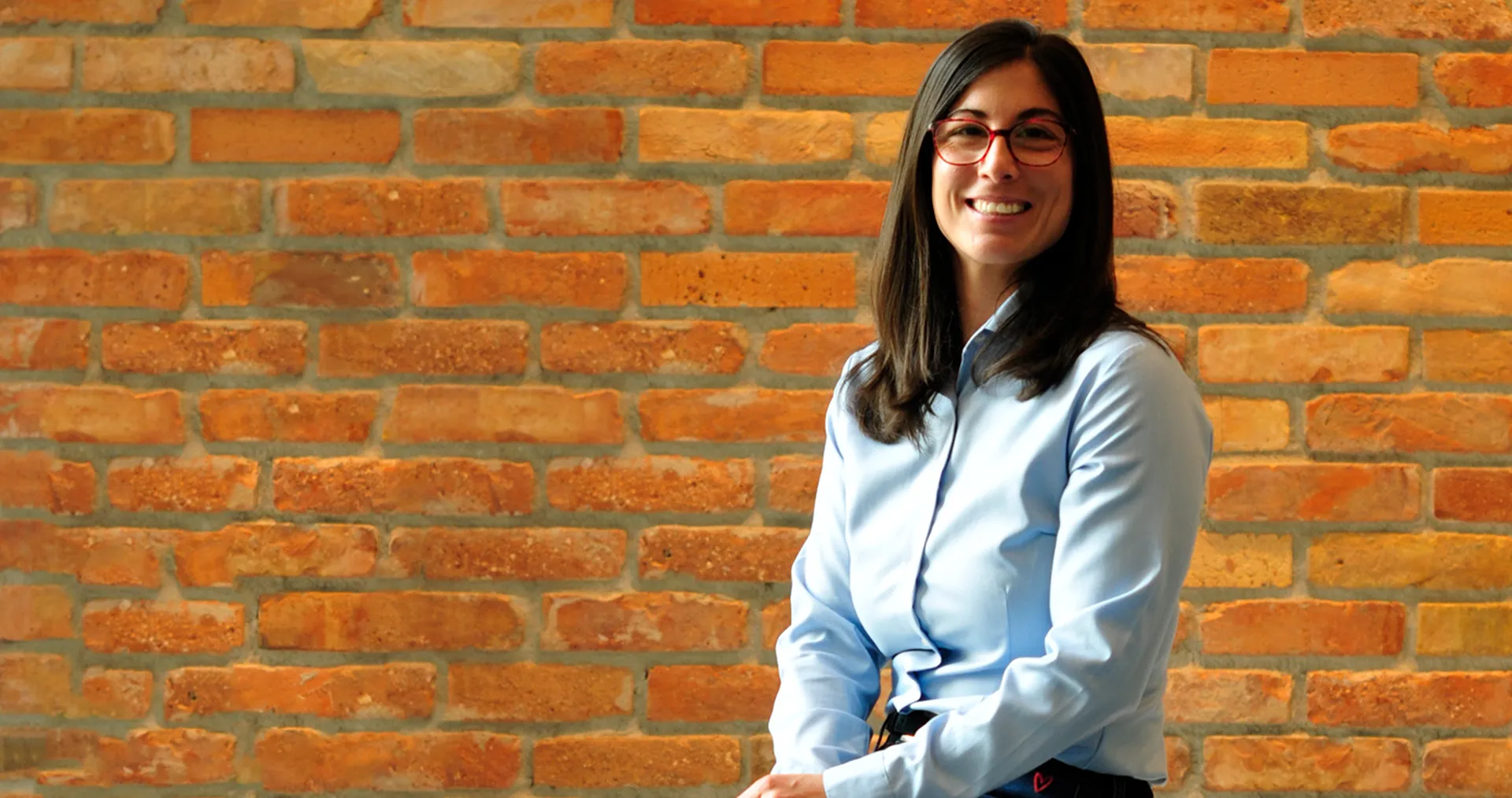 Ágnes Farkas trägt eine Brille und eine blaue Bluse und lächelt vor einer Wand in die Kamera. (Foto)